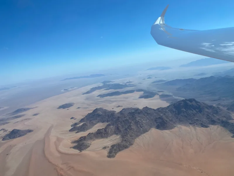 Dark mountains amid pale sands, blue sky and a glider wing on the right